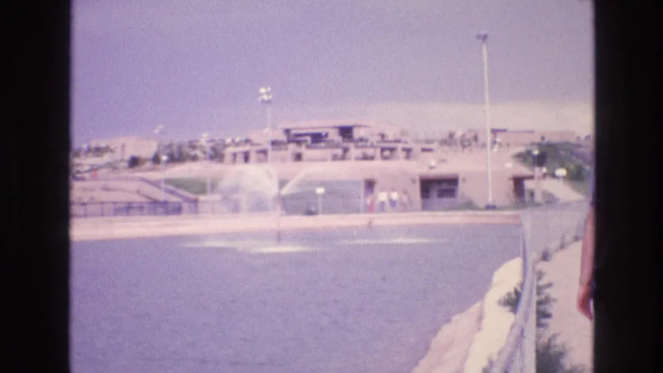 A distant visitor-area view shows a water feature, low buildings, paths, light poles, and small people figures under an overcast sky in New Mexico.