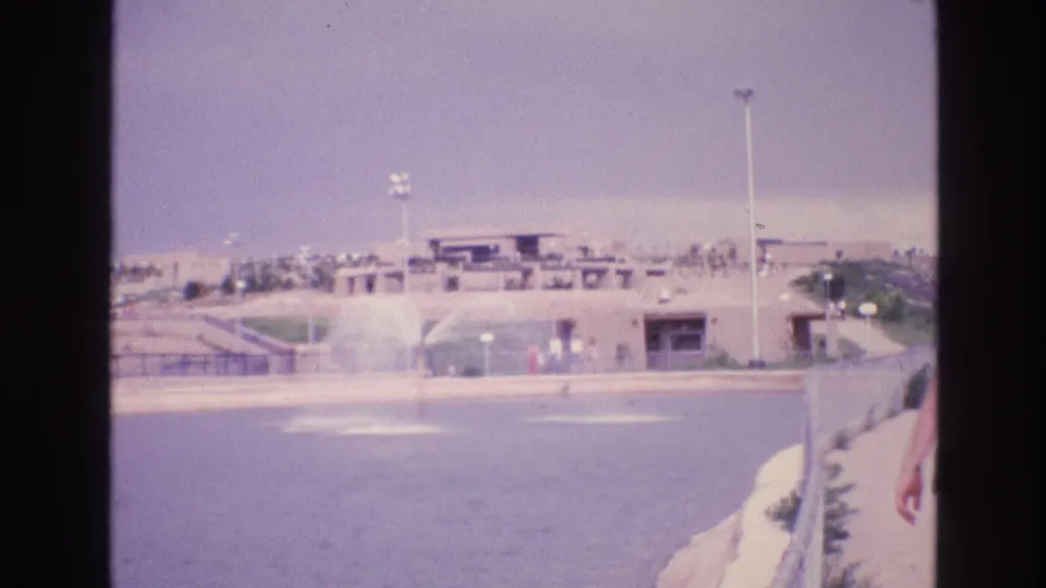 The final frame holds on the wider recreation or zoo area, with a fountain-like water feature, low structures, paths, and distant visitors visible.