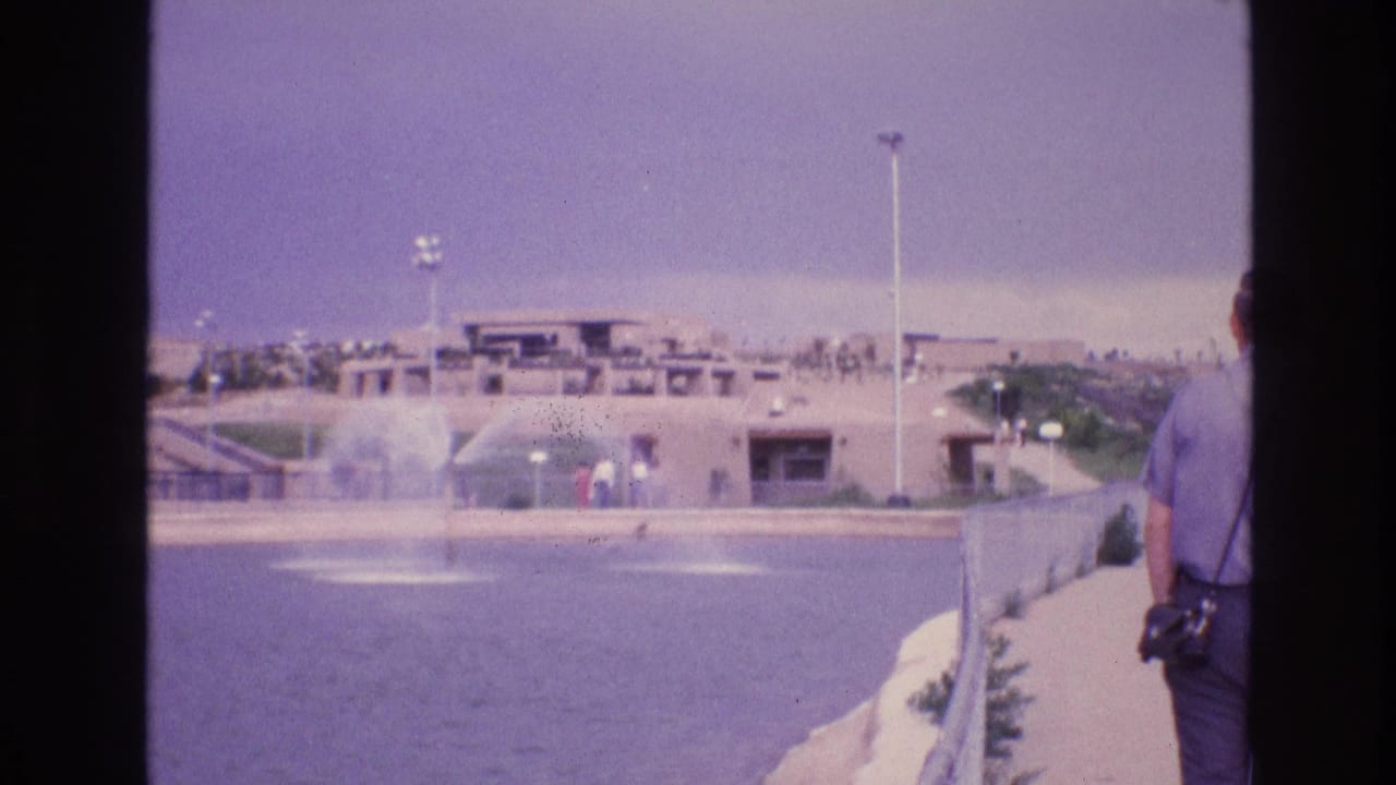 Man with a camera stands near a paved walkway beside fountains and adobe-style buildings.