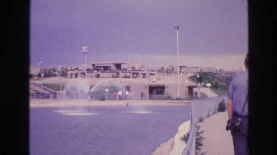 1973 New Mexico home-movie frame showing a man with a camera near a walkway beside an active water feature and adobe-style building complex.