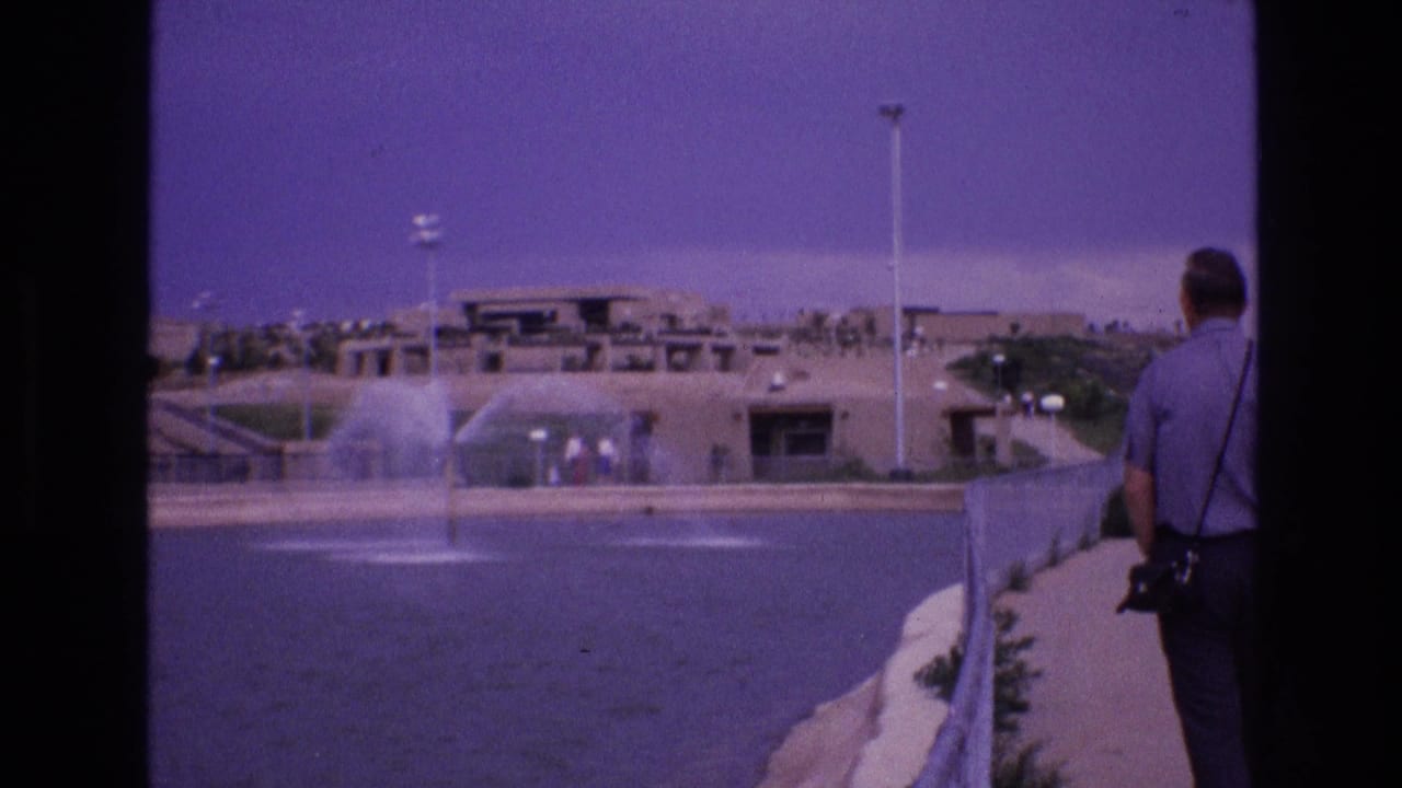 Man stands at the right edge of a walkway looking across a pool with fountains.