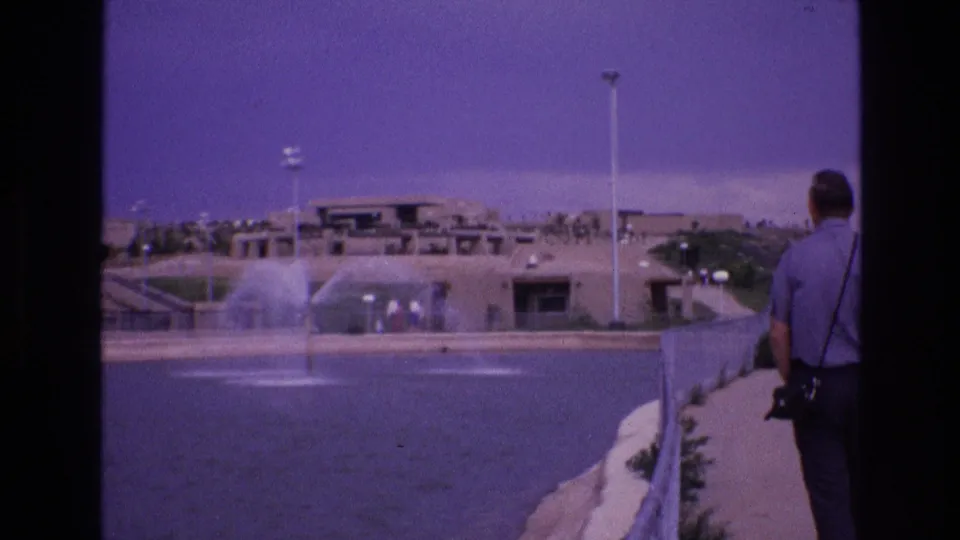 1973 New Mexico travel footage frame with a man at the edge of a paved path looking over a large pool, fountain spray, and low adobe-style structures.