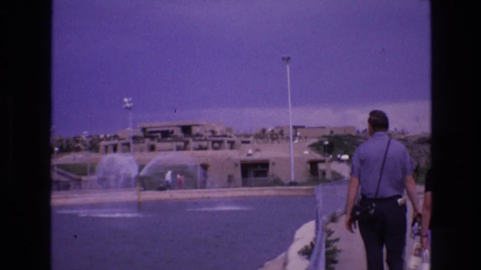 1973 New Mexico 8mm frame showing a male visitor walking along a paved path beside a large water feature, with fountain spray and adobe-style buildings beyond.