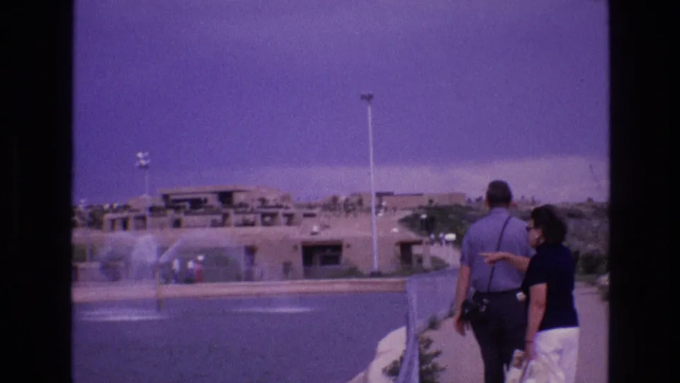 1973 New Mexico home-movie frame showing an adult couple walking along a paved path beside fountains, with low adobe-style buildings and light poles across the water.