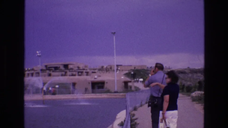 In 1973 New Mexico home-movie footage, a couple seen from behind stands on a walkway near water fountains with low adobe-style buildings beyond.