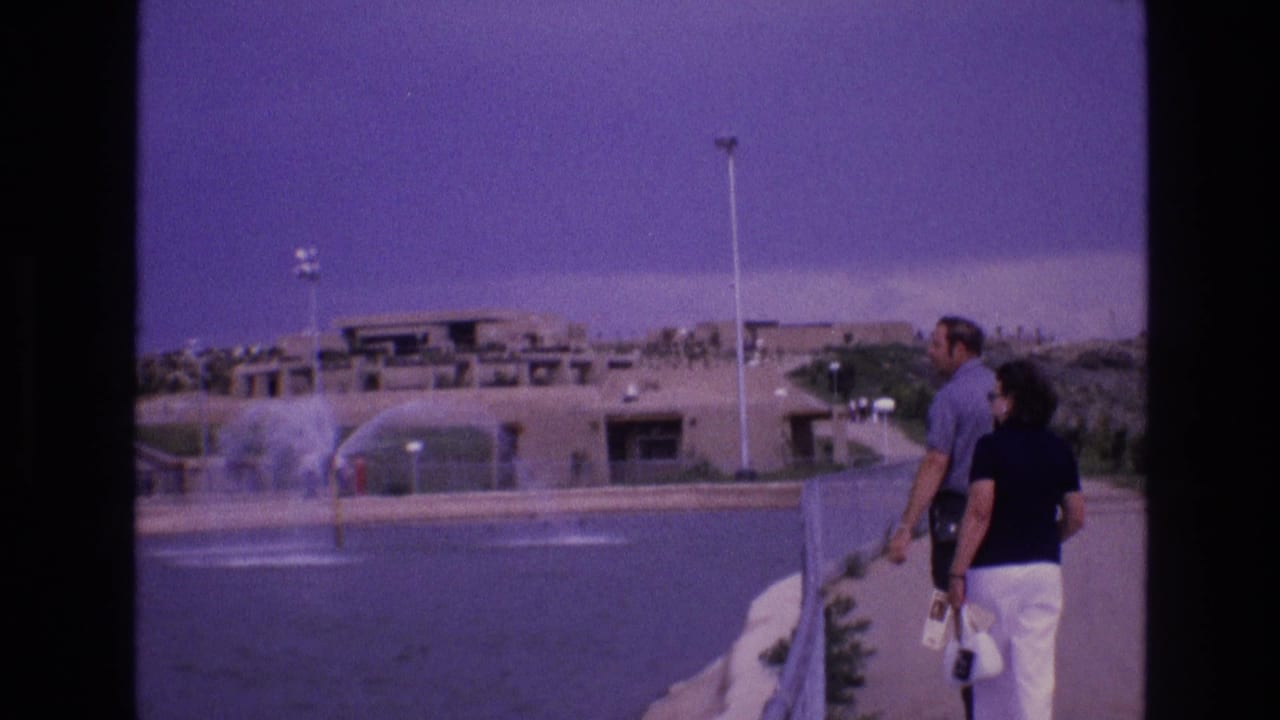 Couple walking along a path beside water fountains and desert landscaping.