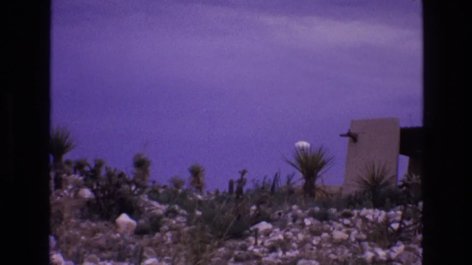 1973 8mm New Mexico desert landscape showing cactus-like plants, rocky terrain, a strong purple film cast, and a partial low wall or structure in the background.