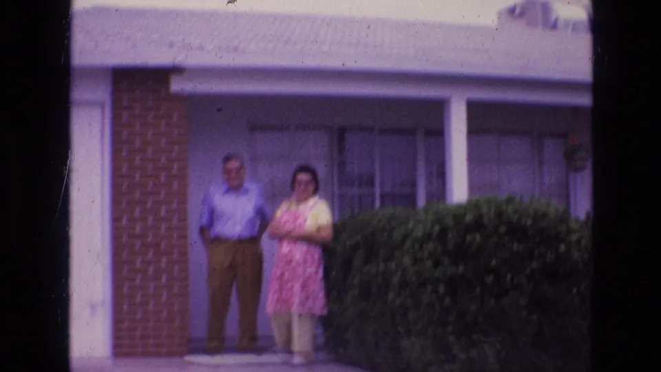 In the supplied 1973 New Mexico home-movie context, a man and woman stand posed on a suburban front porch beside a brick-and-stucco facade and manicured hedge.