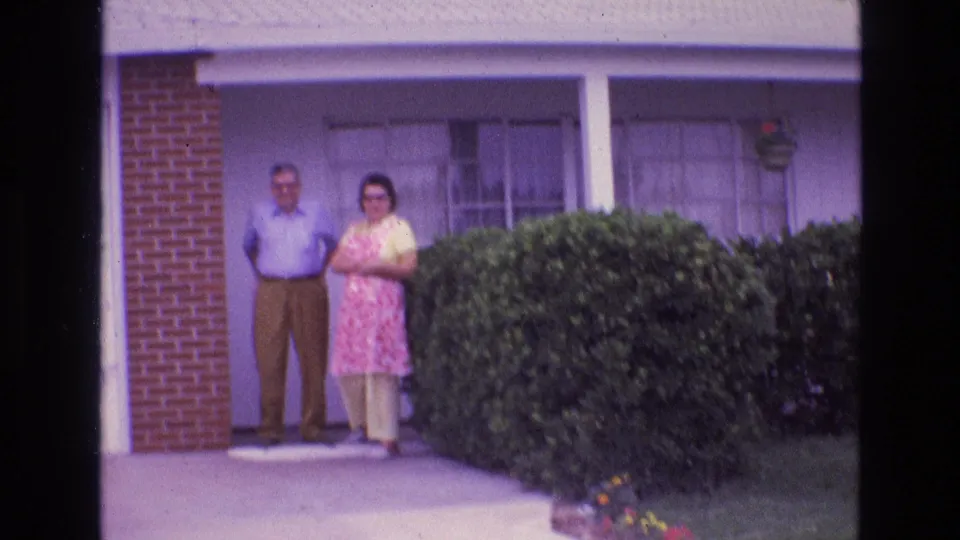 A man and woman stand near the doorway of a single-story suburban house in New Mexico, with porch posts, windows, brickwork, and a trimmed hedge visible.