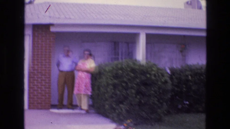 In 1973 New Mexico home-movie footage, a man in a light shirt and a woman in a pink patterned dress stand on the porch of a single-story suburban home.