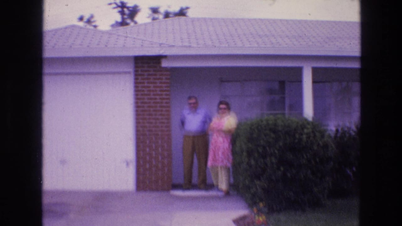 A man and woman stand near a white garage door and front hedge.