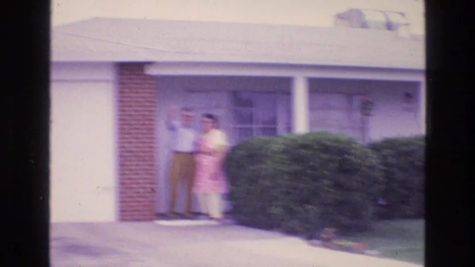 Soft 8mm footage shows the man and woman on the porch of the suburban home, partly framed by a brick pillar, hedge, garage door, and sunlit driveway.