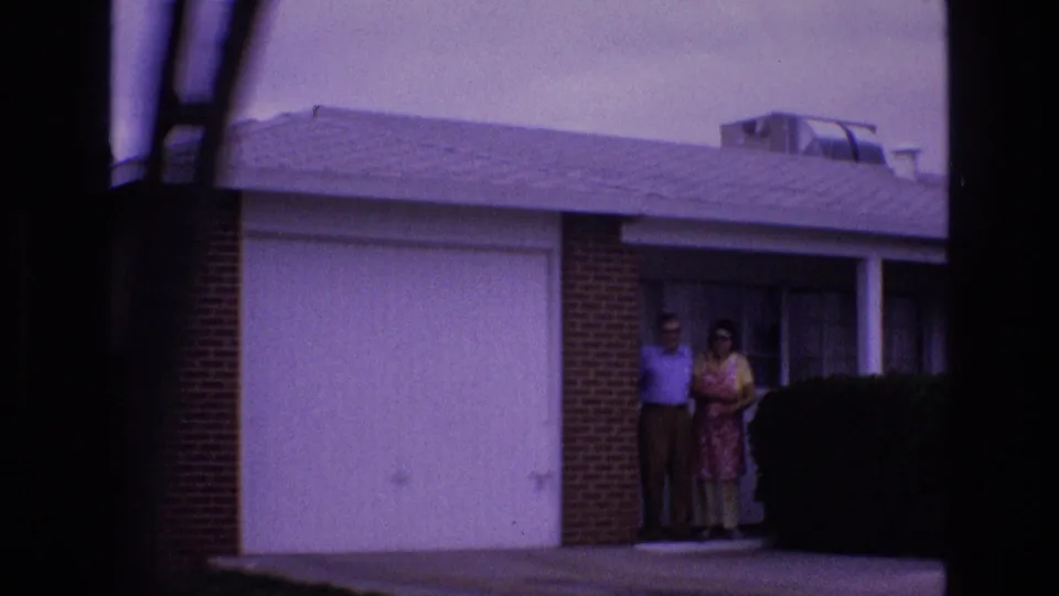 The final frame places the couple deeper in shadow near the porch, with the white garage door, brick facade, dark window, roofline, and hedge defining the suburban exterior.