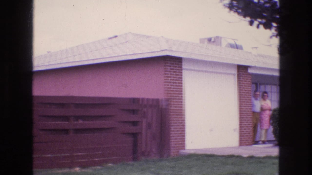 Two adults standing by the porch of a one-story brick and stucco home.
