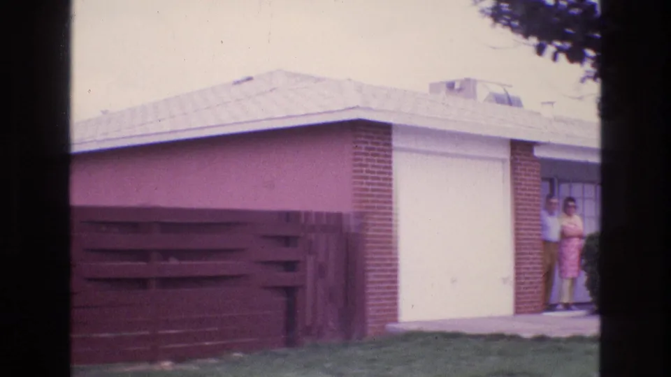 In 1973 New Mexico home-movie footage, two adults stand near a porch doorway beside a one-story brick and stucco house with a wooden fence in front.