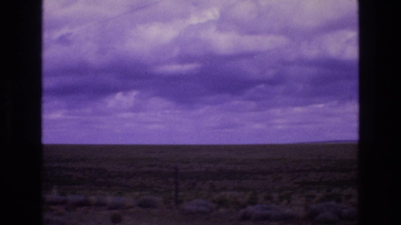 A broad New Mexico plain stretches to the horizon under dense purple-tinted clouds, with low scrub and a fence line visible in the foreground.