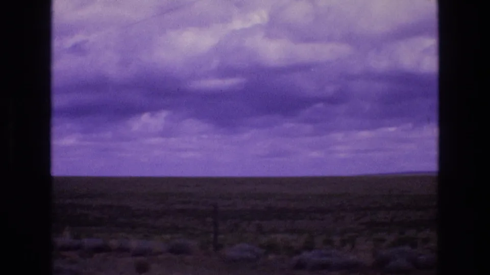 A broad New Mexico plain stretches to the horizon under dense purple-tinted clouds, with low scrub and a fence line visible in the foreground.