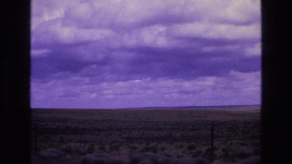 The 8mm frame shows a flat scrubland horizon in New Mexico with a dark fence line and layered clouds across a purple-shifted sky.