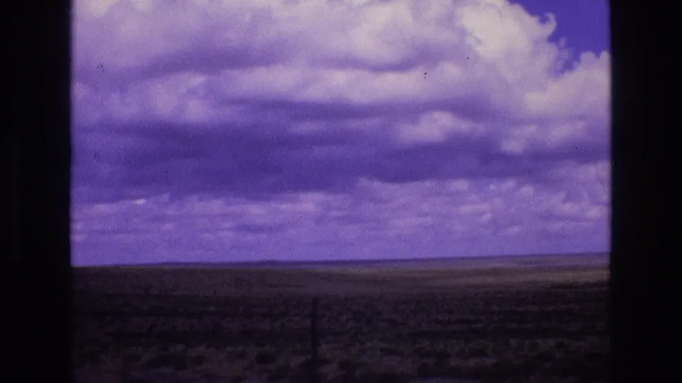 A wide open New Mexico landscape sits beneath large rounded clouds, with the 8mm scan showing strong purple color shift and soft distant detail.