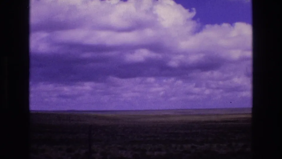 1973 New Mexico 8mm home-movie frame showing open grasslands under a purple-blue sky with cumulus clouds and visible film gate borders.