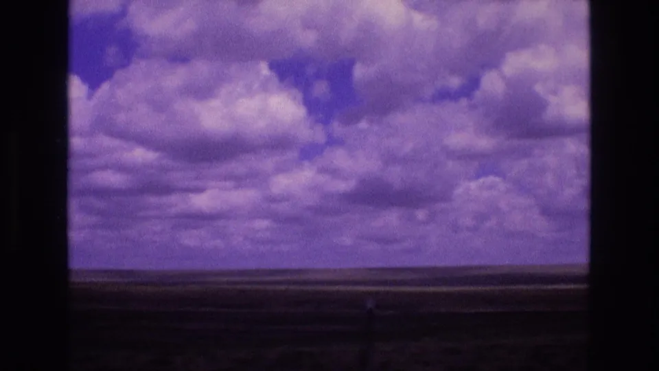 1973 New Mexico home-movie frame showing purple-tinted cumulus clouds over open grasslands with visible 8mm borders and analog grain.