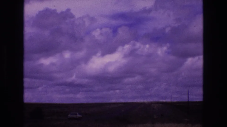 1973 New Mexico 8mm frame showing layered clouds, open land, window framing, and a small light-colored vehicle-like shape near the lower horizon.