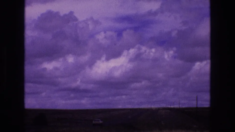 1973 New Mexico 8mm frame showing a dark desert horizon, stormy purple sky, a small highway vehicle, and utility poles in the distance.