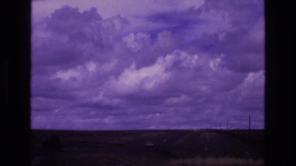 1973 New Mexico road-travel frame with a broad clouded sky, low desert landscape, distant vehicle shapes, utility poles, and a roadside embankment.