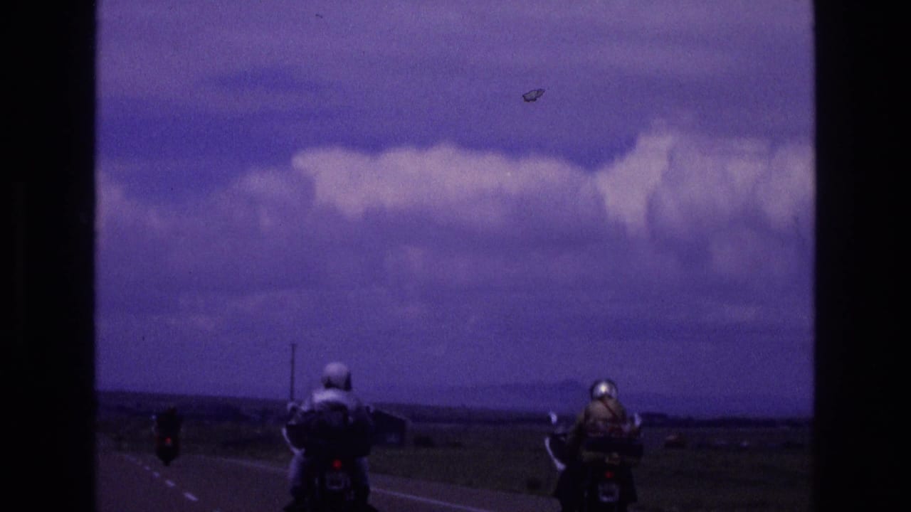 Two helmeted motorcyclists ride away on a rural two-lane road beneath a clouded sky.