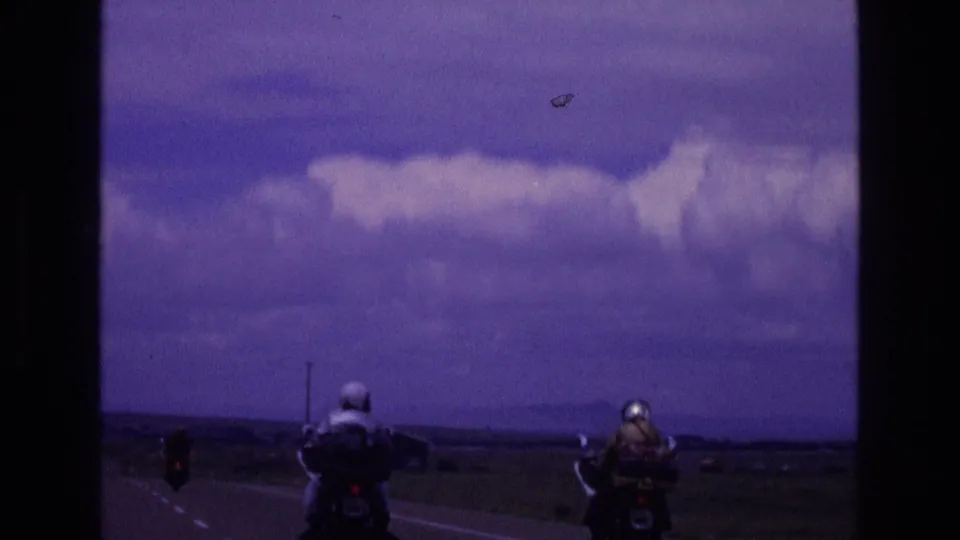 In 1973 New Mexico home-movie footage, two helmeted motorcyclists ride away along a rural two-lane highway under a purple-tinged sky with large clouds.