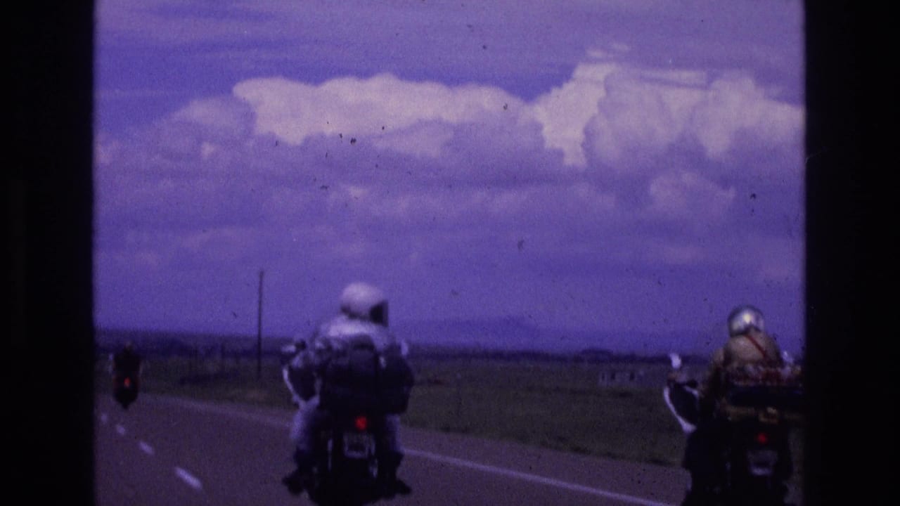 Two motorcyclists continue along an open highway with fields and clouds in the distance.