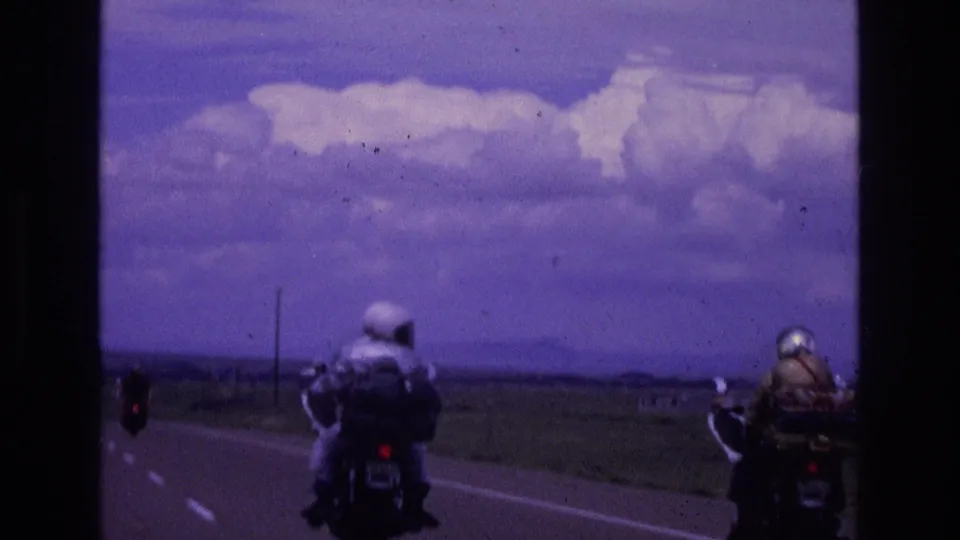 Two helmeted riders continue down a rural New Mexico highway in 1973 footage, seen from behind with open land, distant hills, and a cloud-filled sky ahead.