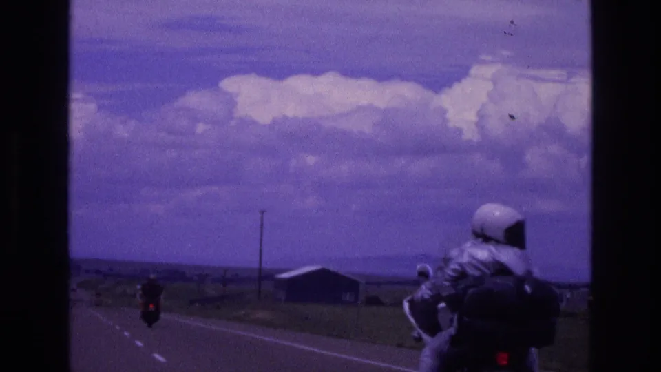 A trailing 8mm view in New Mexico shows a helmeted motorcyclist closer to the camera as another rider and a small roadside building appear along the rural highway.