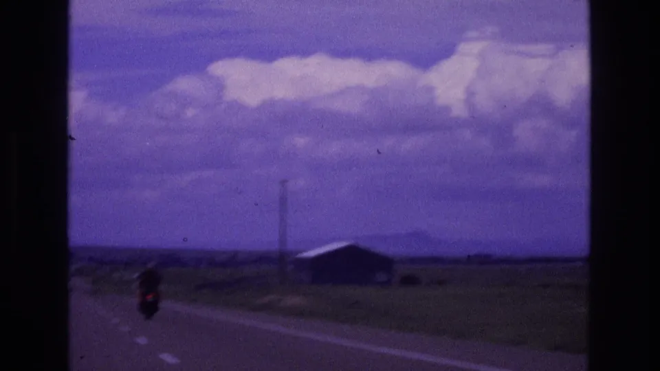 A lone motorcyclist rides away on a rural New Mexico road in 1973 footage, passing open fields, a small building, utility poles, and distant low hills beneath large clouds.