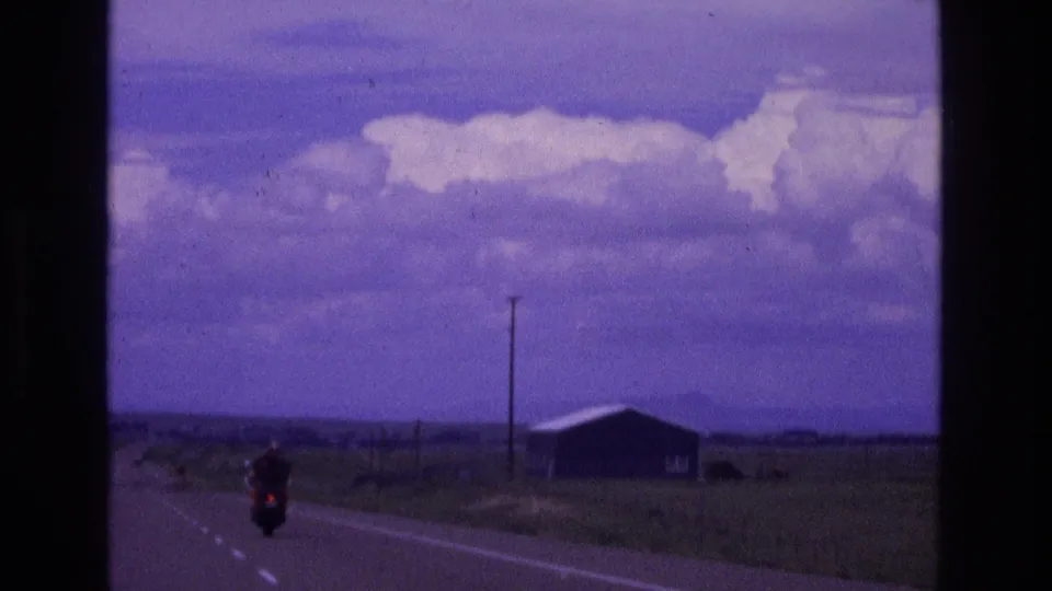 A lone motorcyclist rides along a rural New Mexico highway in 1973, with open land, a small roadside building, and a utility pole visible under a purple-shifted sky.