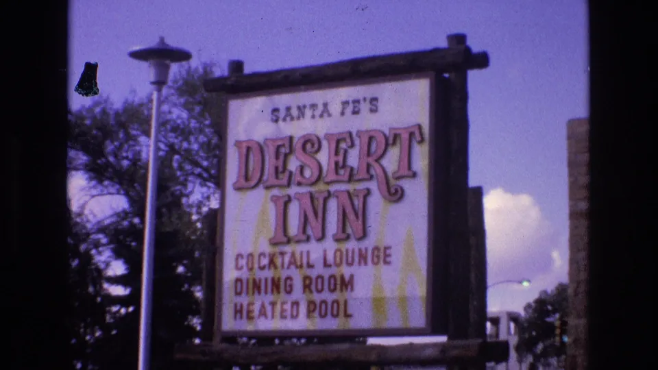 1973 home-movie frame from Santa Fe, New Mexico showing the Desert Inn roadside sign with cocktail lounge, dining room, and heated pool text, framed by trees and a street lamp.