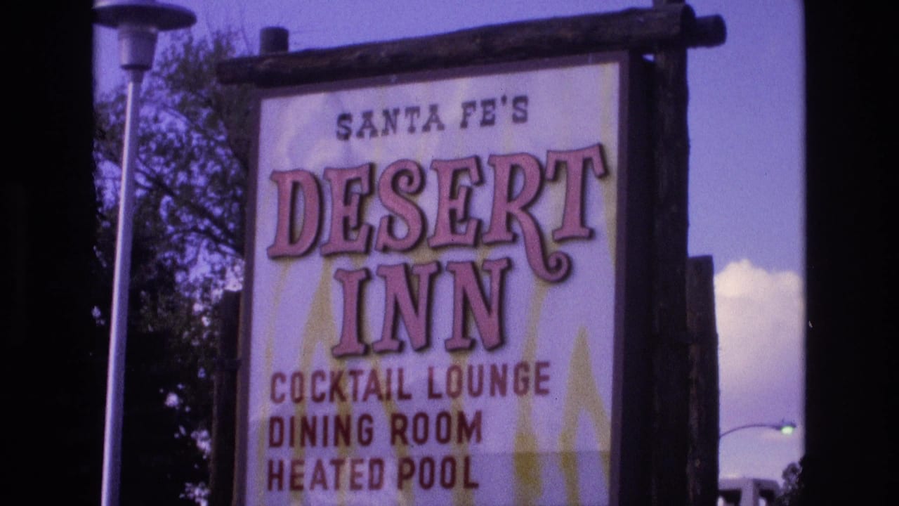 1973 Santa Fe, New Mexico frame showing a closer view of the Desert Inn sign with pink lettering, amenity text, wooden supports, trees, and blue-purple sky.