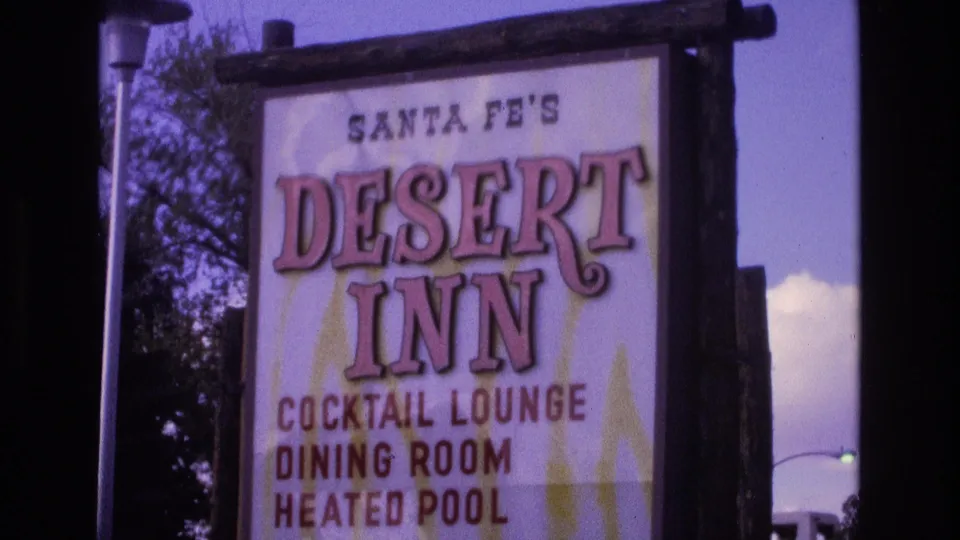 Final 1973 Santa Fe frame showing the Desert Inn roadside sign in a tight home-movie composition, with wooden framing, amenity lettering, trees, and purple-shifted sky.