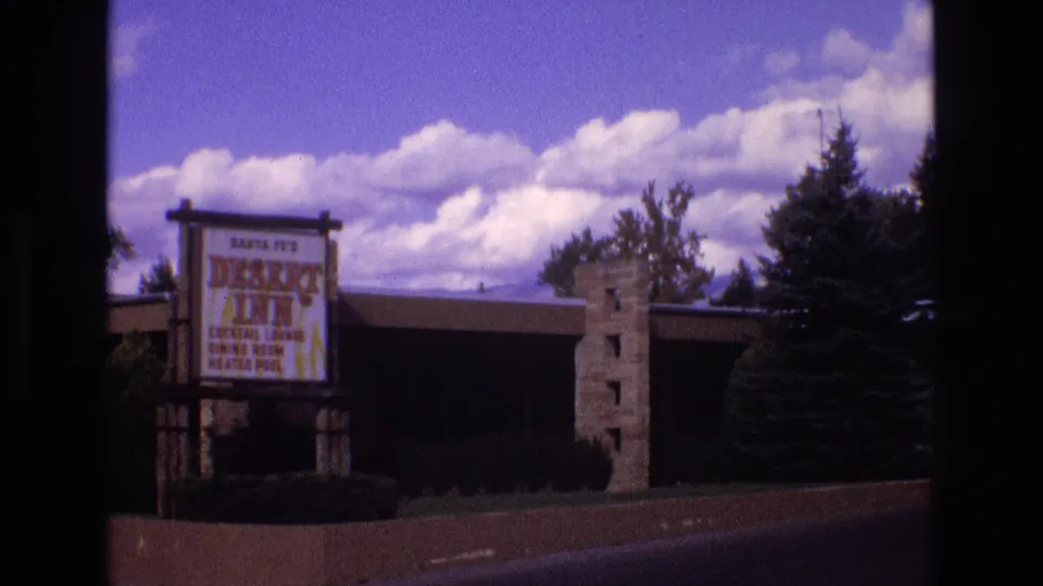 Wider 1973 Santa Fe view showing the Desert Inn roadside sign, low motel building, stone pillar, shrubs, trees, and a cloudy blue sky.