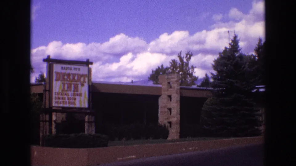 Wide 1973 New Mexico 8mm frame with the Desert Inn sign at left, motel facade, stone entry pillar, dark trees, and large white clouds overhead.