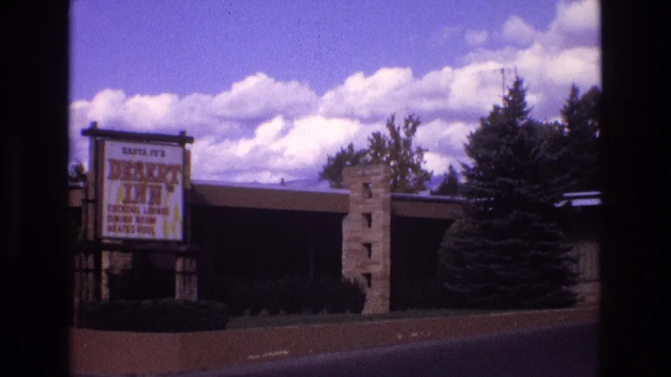 1973 New Mexico home-movie frame showing the Santa Fe’s Desert Inn sign beside a low motel building, trees, roadway, and clouded sky.