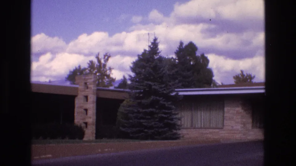 1973 New Mexico home-movie frame showing the motel facade, stone column, evergreen tree, curtained windows, road surface, and cloud-filled sky.