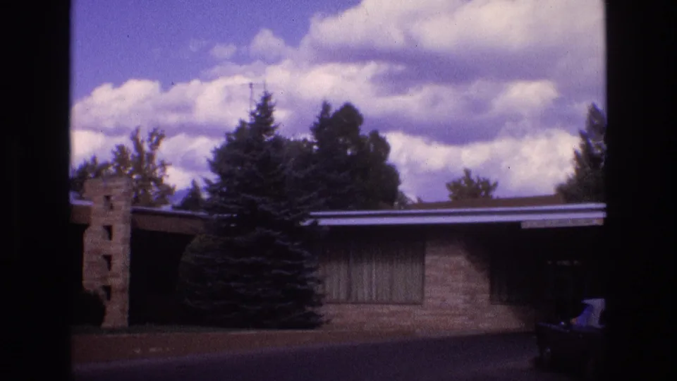 1973 New Mexico frame showing the motel’s low facade, stone pillar, evergreen tree, roadway, clouded sky, and a dark car shape entering the lower-right edge.