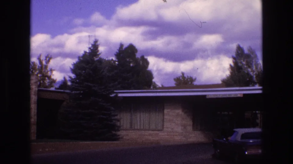 1973 New Mexico 8mm frame showing a low mid-century-style brick house with a flat roof, evergreen trees, dramatic clouds, and part of a parked car in the driveway.