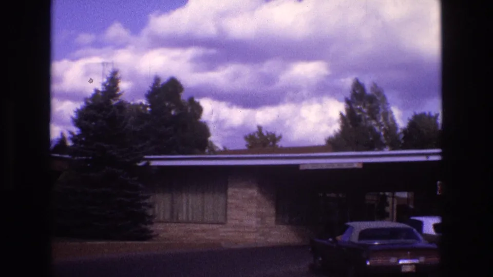 1973 New Mexico home-movie frame of a single-story brick residence with a flat roofline, trees behind it, and a dark car parked near the front.
