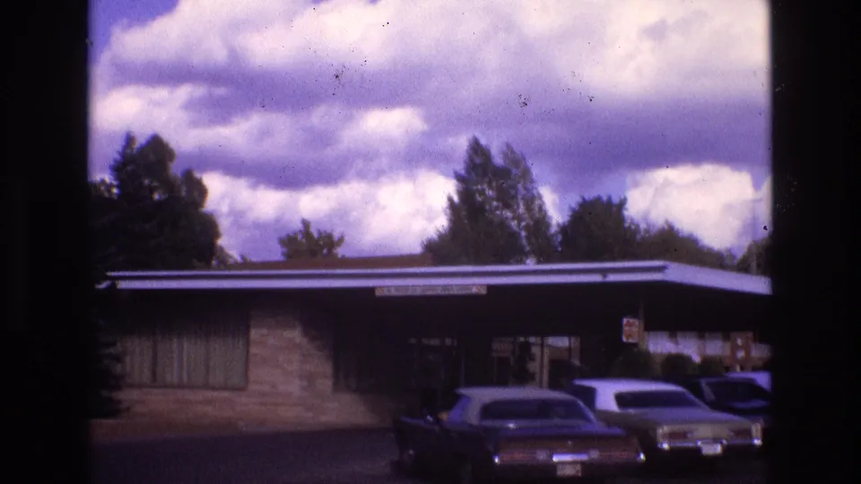1973 New Mexico 8mm frame showing the camera pan across a brick residential exterior with a flat roof, broad clouds, and multiple parked cars in the driveway.