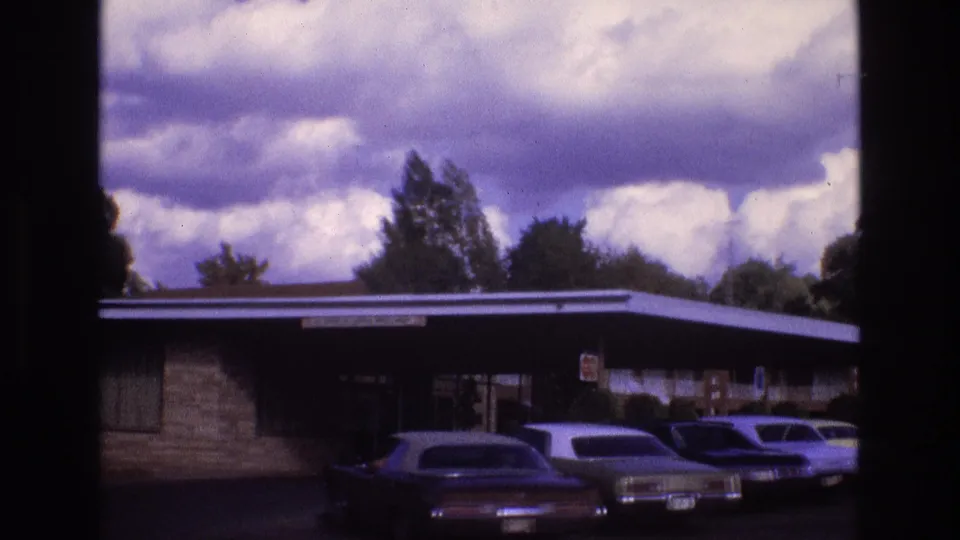 1973 New Mexico home-movie frame showing a low brick house with a flat roof and several period cars parked across the driveway beneath a cloud-filled sky.