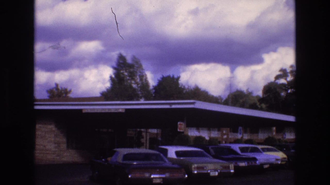 1973 New Mexico 8mm frame showing a low roadside motel exterior with several parked vintage cars beneath a dramatic cloud-filled sky.