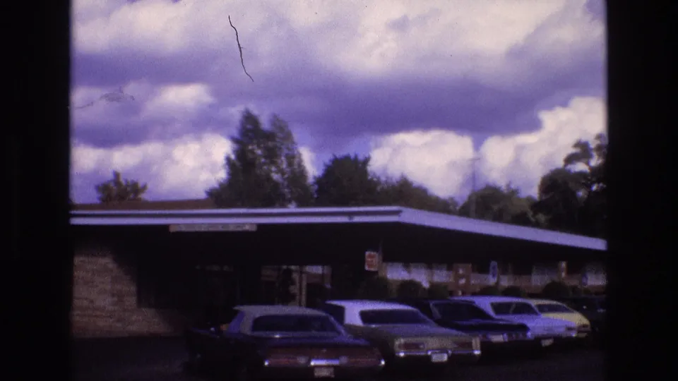 1973 New Mexico 8mm frame showing a low roadside motel exterior with several parked vintage cars beneath a dramatic cloud-filled sky.