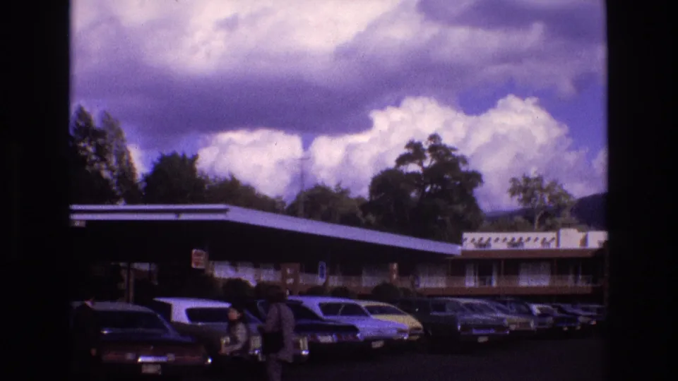 1973 New Mexico roadside motel scene with rows of vintage cars and people walking through the parking lot in front of the building.